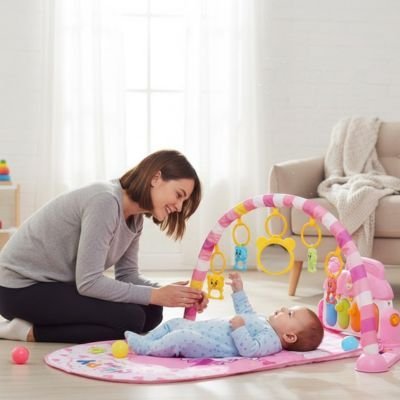 Arche d'éveil bébé avec un tapis moelleux rose, présentée par une maman jouant avec son bébé allongé. Jouets suspendus colorés.