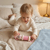 Jouet musical bébé en rose avec motifs souriants, manipulé par un enfant allongé sur un lit avec des draps clairs.